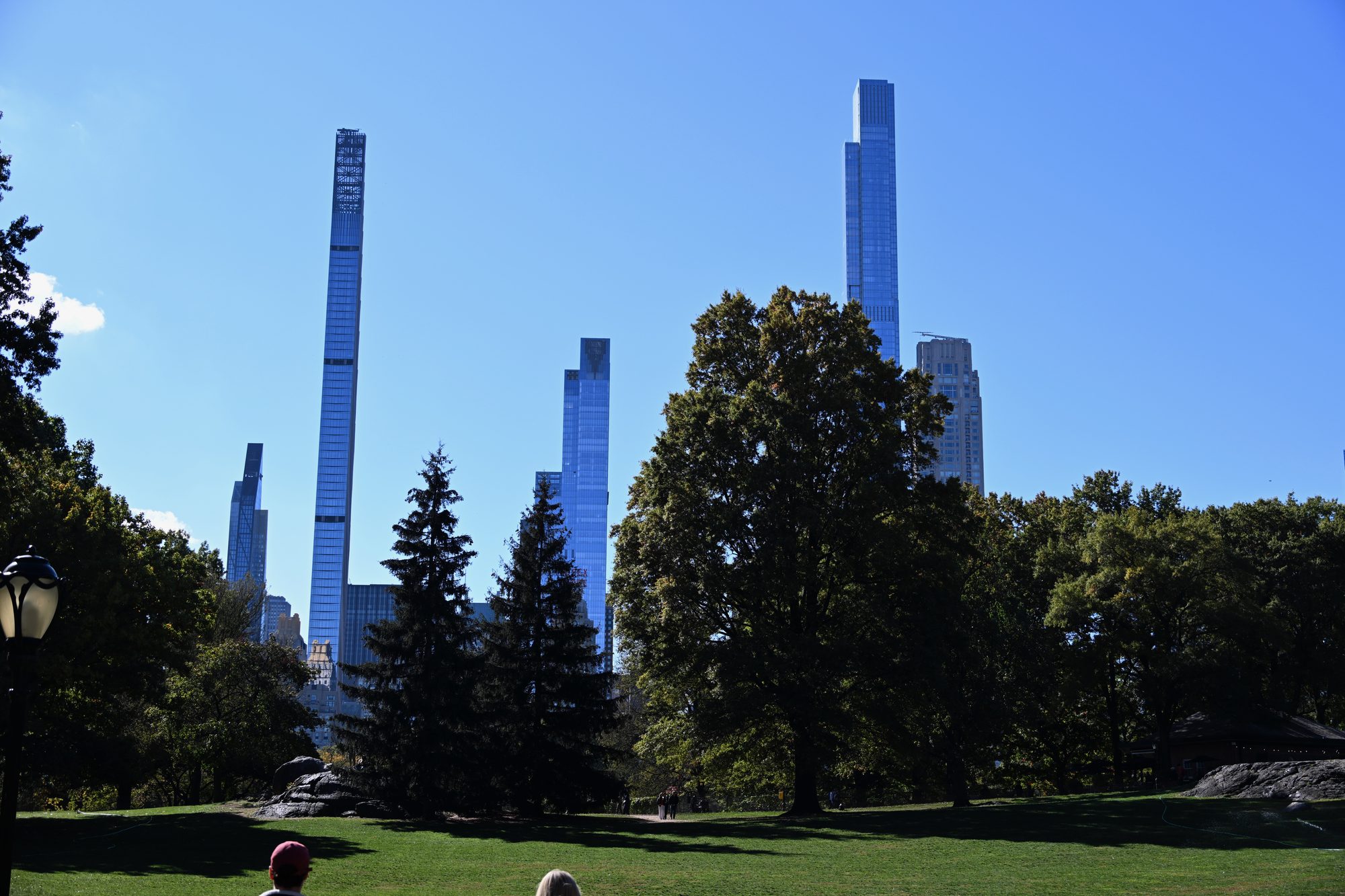 City skyline behind a park with lush green trees and a calm body of water in the foreground under a partly cloudy sky.