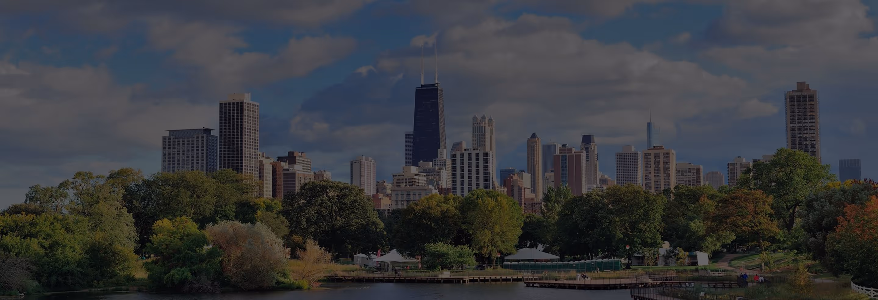 City skyline behind a park with lush green trees and a calm body of water in the foreground under a partly cloudy sky.