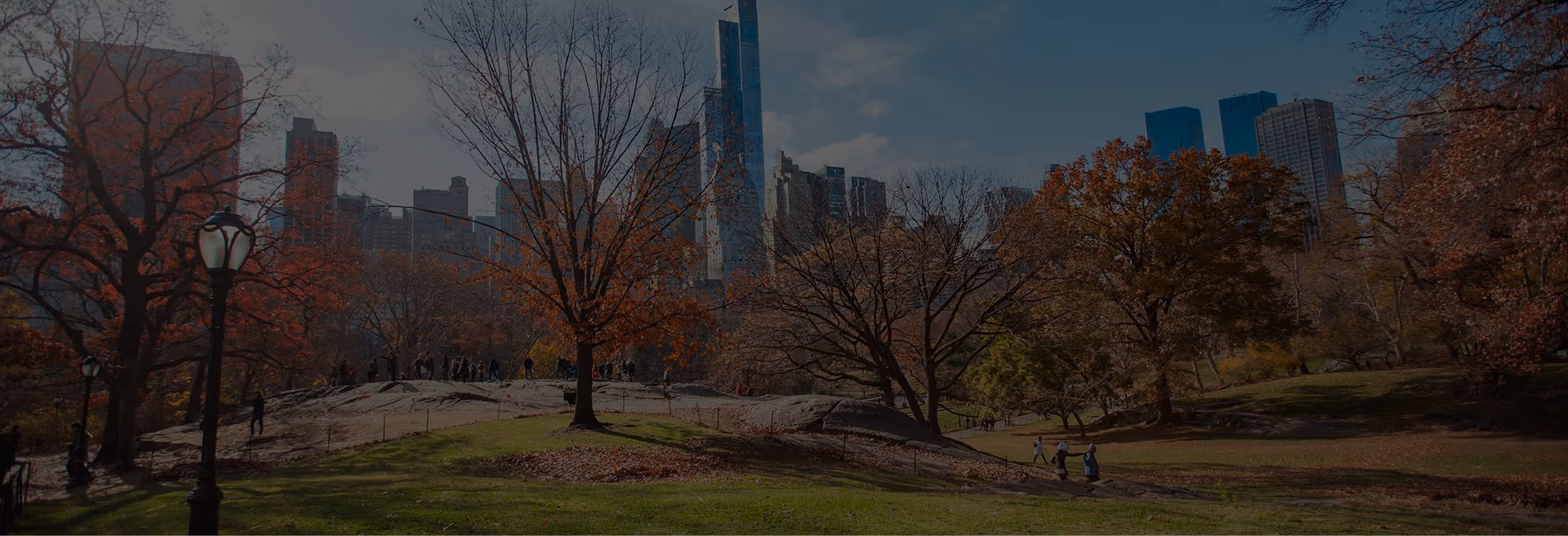 Autumn scene in an urban park with people walking among trees and skyscrapers in the background.